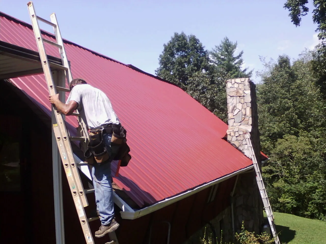 Roofer on a ladder beside a red metal roof and stone chimney — Soffit & Fascia Repair by Roofing Brothers in Readsboro